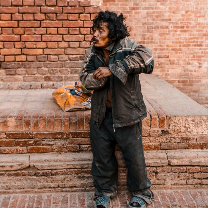 A man in worn clothing standing against a brick wall, reflecting on public horror stories and life's hardships.