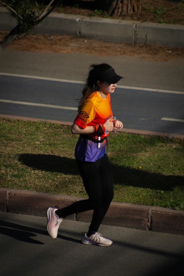 A female runner in a colorful shirt and black visor runs along a paved road next to a grassy median, showing an unusual way people lost lives.