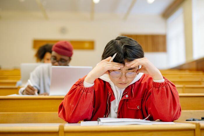 A U.S. student in a red jacket, wearing glasses and looking stressed during an exam, with another student in the background. She is likely taking a test to determine if she is smarter than the average U.S. student. A U.S. student in a red jacket, wearing glasses and looking stressed during an exam, with another student in the background. She is likely taking a test to determine if she is smarter than the average U.S. student.