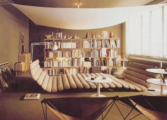 A living room with an iconic furniture piece, a long, low, rippled beige lounge chair, facing bookshelves, under a suspended white fabric ceiling.