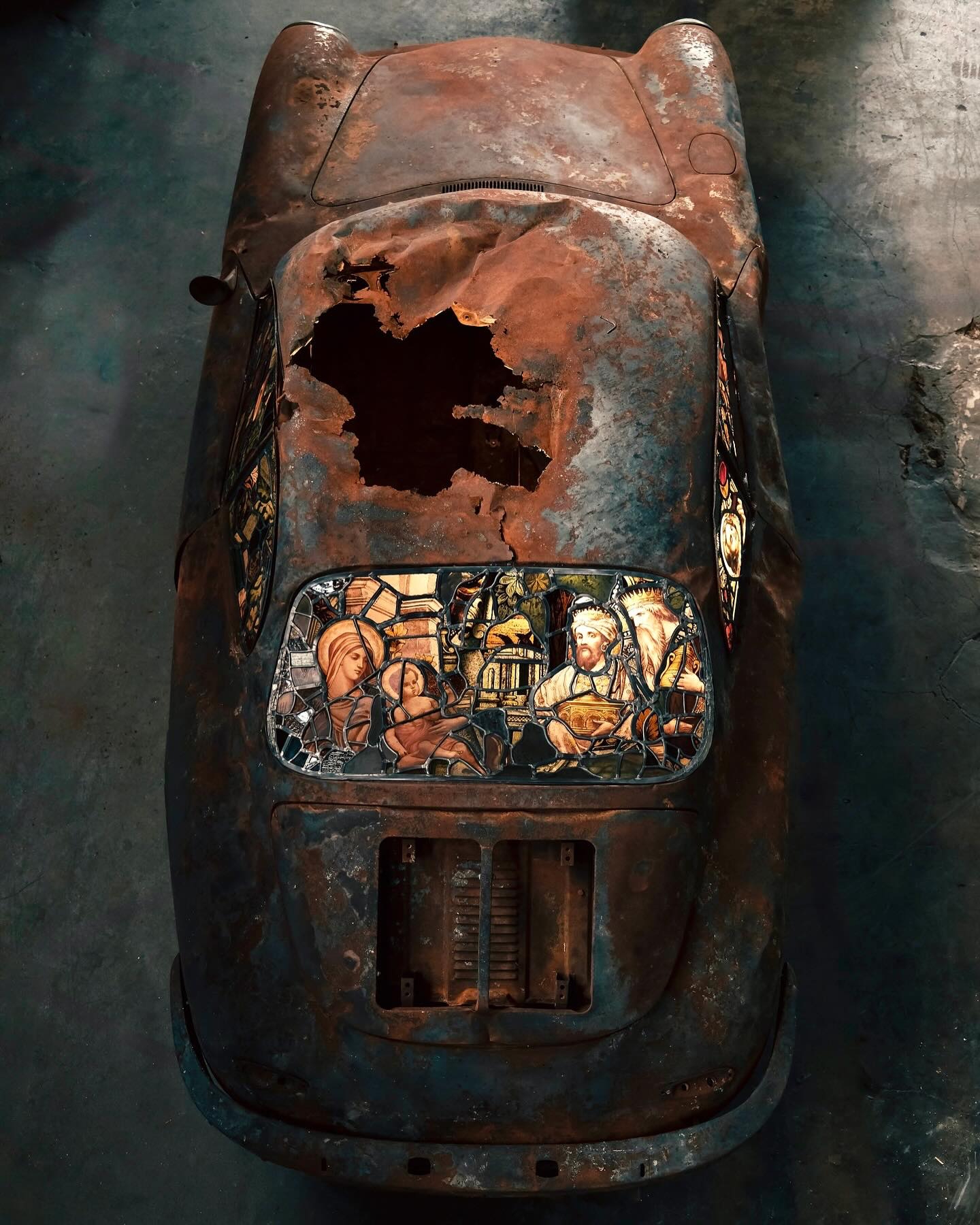 Overhead view of a burned and rusted Porsche 911 with an extraordinary stained-glass sculpture rear window.
