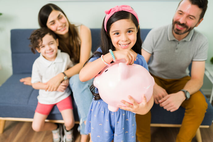A young girl happily puts money into a piggy bank, with her family in the background, demonstrating house etiquette.
