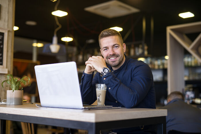 Smiling man with a beard sitting at a table with a laptop and a drink, representing jobs guys have.