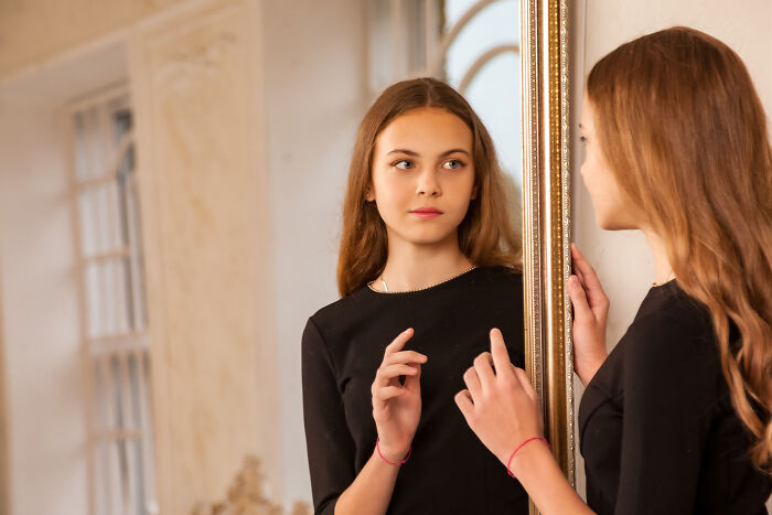 A young woman in a black top gazes at her reflection in a gold-framed mirror, revealing interesting facts about self-perception.