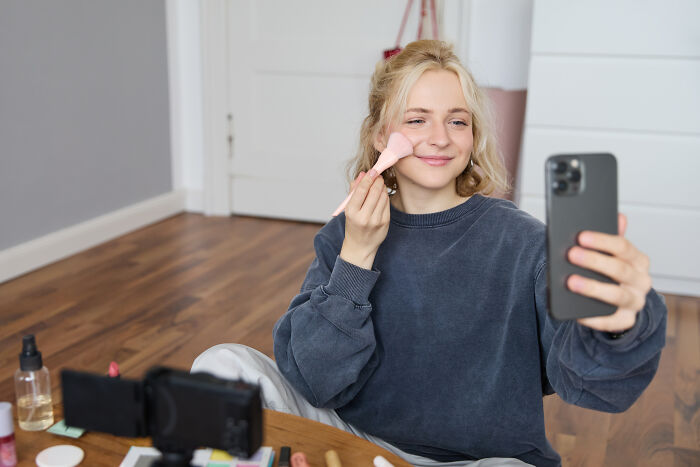 A young woman applying makeup with a brush while holding a phone, demonstrating guests ignoring house etiquette.