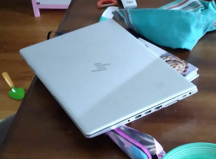 A silver laptop with an HP logo rests on a wooden table, partially obscuring a book and a colorful pouch, a real photo that makes your brain question if it was edited.