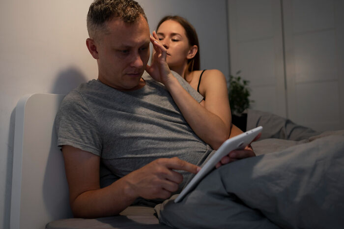 A man on a tablet in bed, ignoring red flags of a sociopath, while a woman whispers to him.