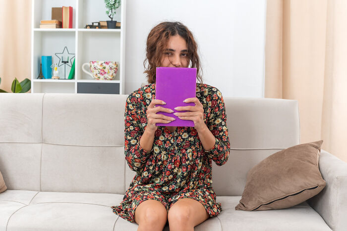 Woman in a floral dress on a couch, holding a book to her face. Ignoring red flags because the person was hot.