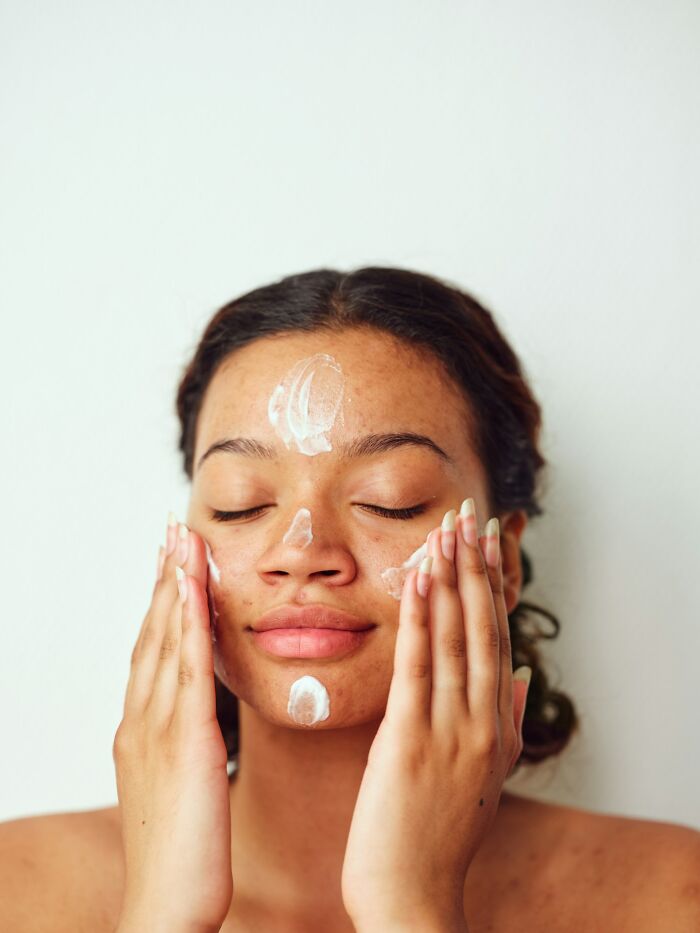 Woman applying face cream with dots on her forehead, nose, and chin, revealing companies' beauty secrets.