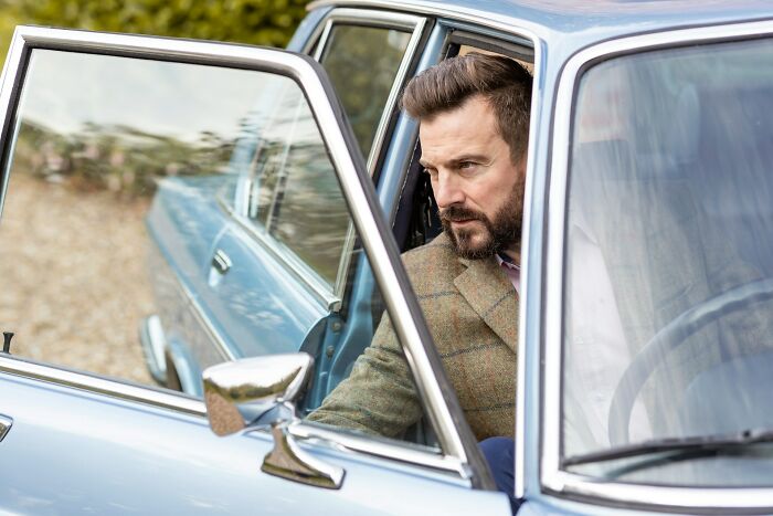 A man with a beard looking intently from inside a car with the door open, showing a serious 'we need to leave now' expression.