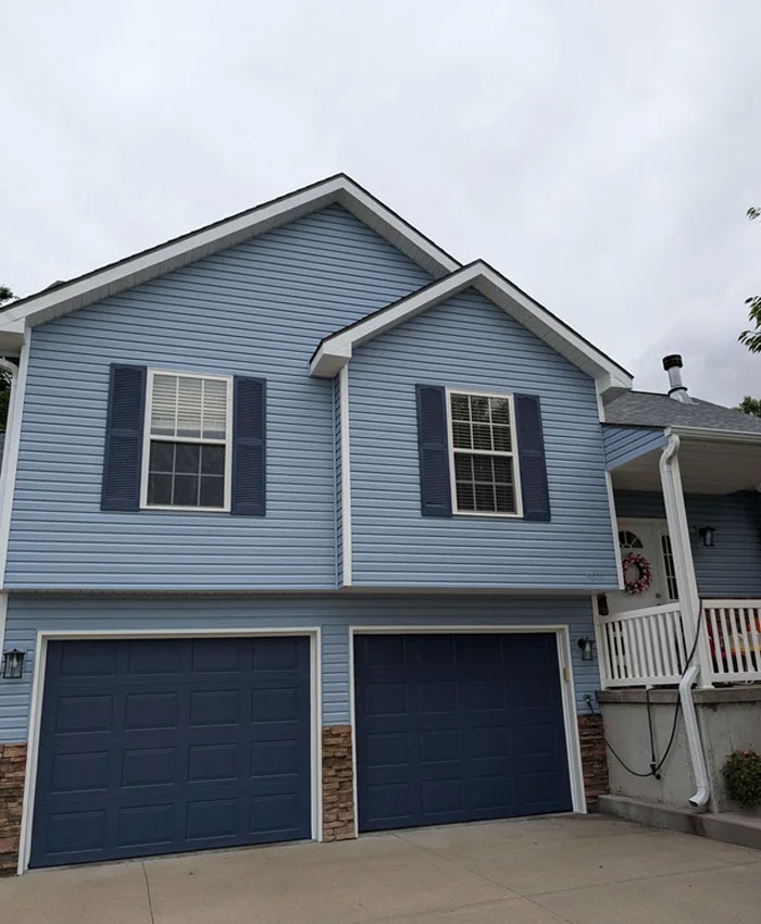 A two-story blue house with dark blue garage doors, representing people asking for help fixing things related to home exteriors.