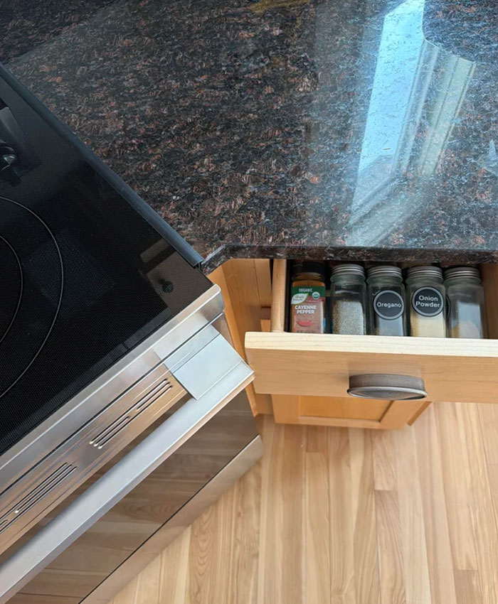 People ask others for help fixing an issue with a kitchen drawer next to a stove, filled with spice jars.