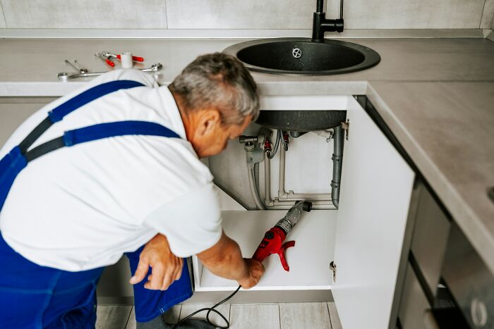 A plumber working on pipes under a sink, showing how people are getting scammed by dishonest repair services.