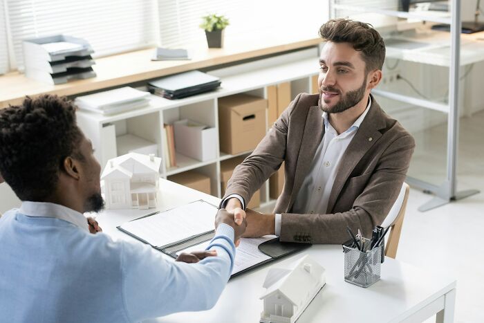 Two men shaking hands across a desk with small house models, showing how people are getting scammed in transactions.