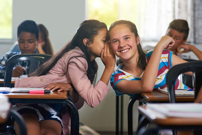 Two girls in a classroom; one whispers to the other, who smiles at the camera. Teachers claim they can spot red flag students.