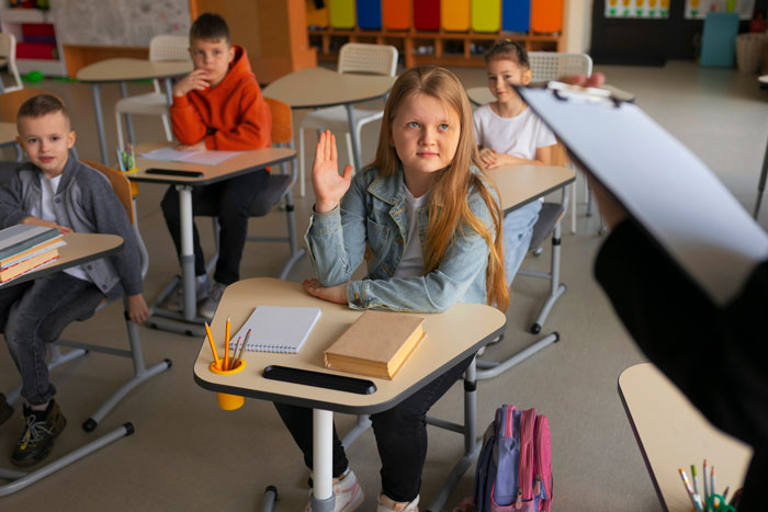 A young girl in a classroom raising her hand, with other students and a teacher visible. Teachers claim they can spot red flag students.