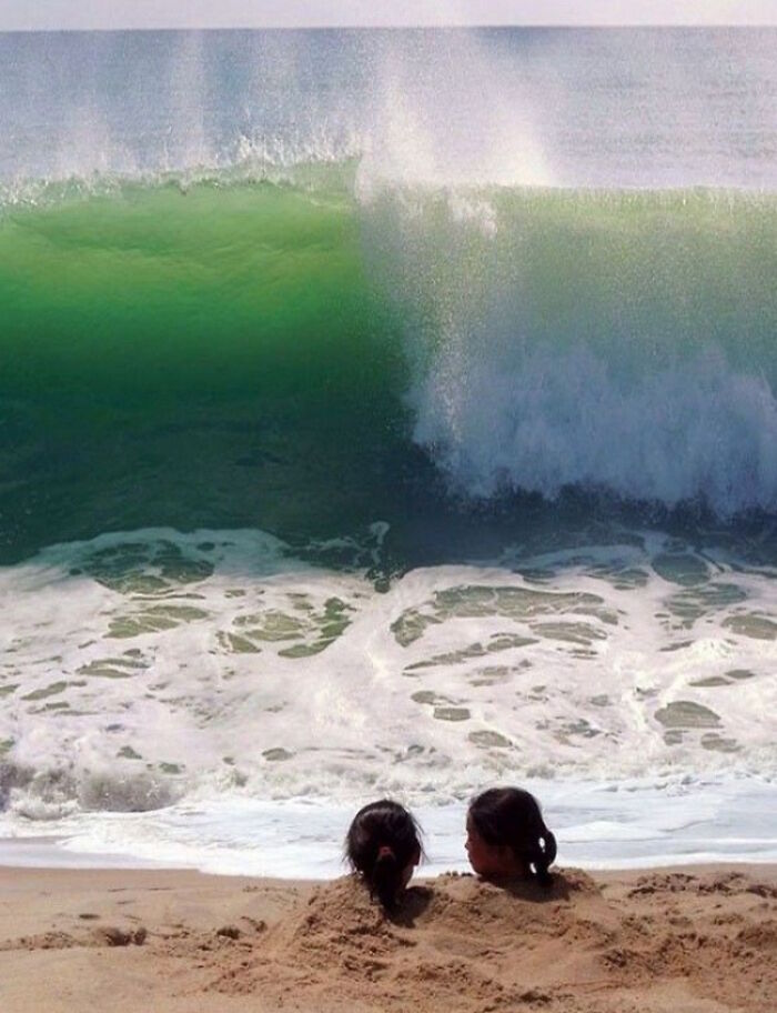 Two heads buried in sand face a massive wave, a hilarious photo taken moments before disaster strikes.