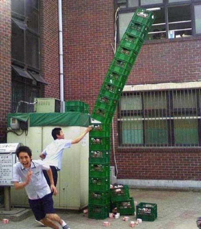 Two men caught in hilarious photos, attempting to balance a tall stack of green crates moments before disaster.