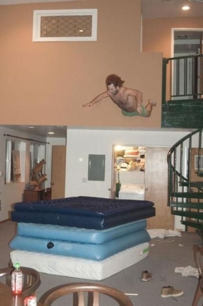 A cutout of a man is placed high on a wall, above stacked air mattresses, depicting a moment before disaster.