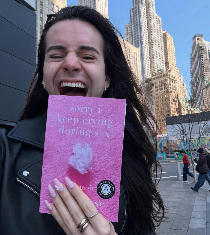 Trans influencer smiling, holding a pink book, with city buildings in background, relevant to Lindsey Graham's hotel claims.