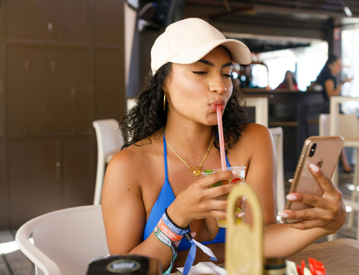Travel influencer taking a selfie while drinking, wearing a blue bikini and cap, lying about solo trips. Travel influencer taking a selfie while drinking, wearing a blue bikini and cap, lying about solo trips.