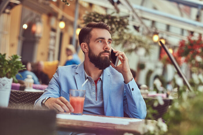 A bearded man in a blue jacket sits at an outdoor cafe, talking on his phone, representing psycho moments from partners.