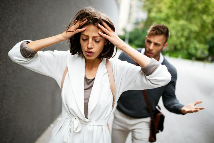 A woman looking stressed, holding her head, with a man arguing behind her, depicting psycho moments from partners.