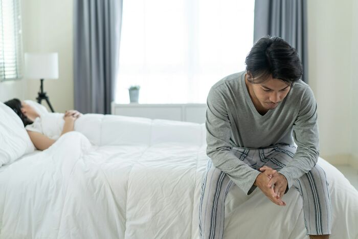 A man sitting on the edge of a bed, looking down sadly, with a woman lying in the background, showing psycho moments from partners.