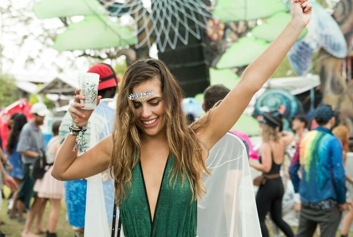 A woman with embellished face paint and a green outfit dances happily, holding a drink, at a lively event, celebrating psycho moments.