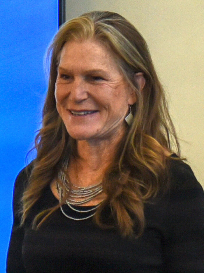 Smiling woman with long brown hair, wearing a black top and layered necklace, shares cool history facts.