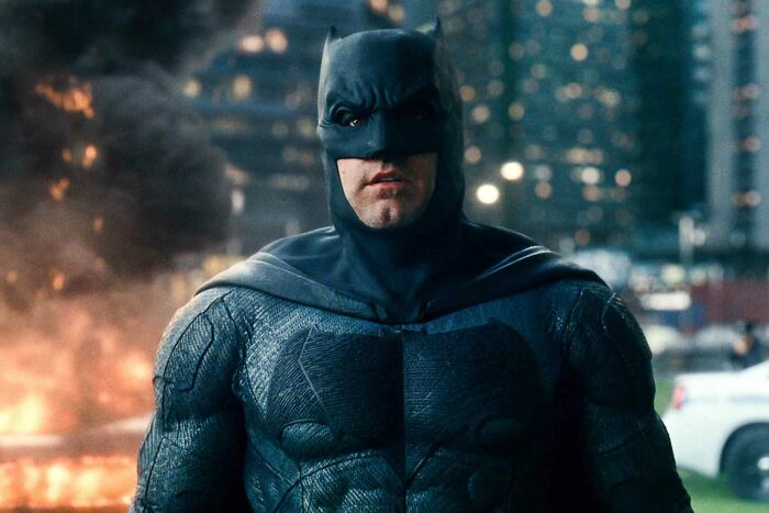 Batman in costume against a backdrop of smoke and city lights, a cool history fact about superhero cinema.