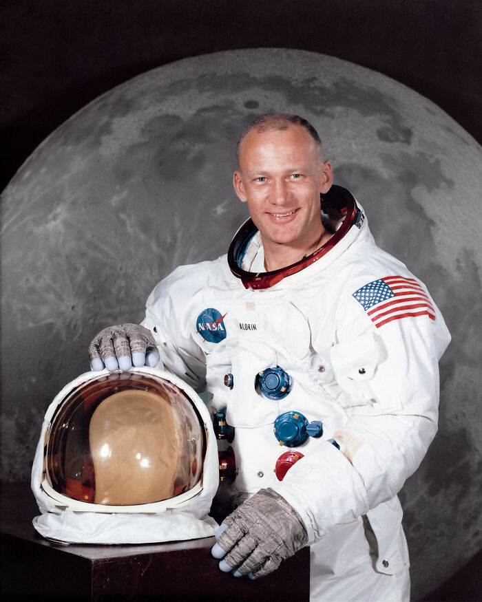 Buzz Aldrin in a spacesuit, smiling, holding his helmet with a moon background, revealing coolest history facts.