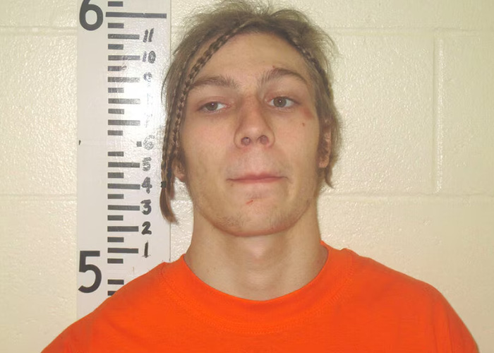 Mugshot of a young man with braids and an orange shirt, standing against a height chart, involved in a sibling dispute.