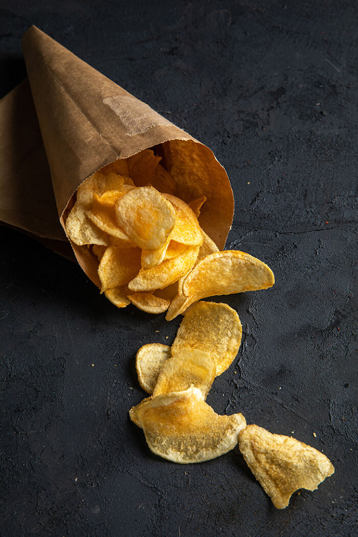 Crispy potato chips spill from a brown paper cone onto a dark surface, representing interesting facts about snack foods.