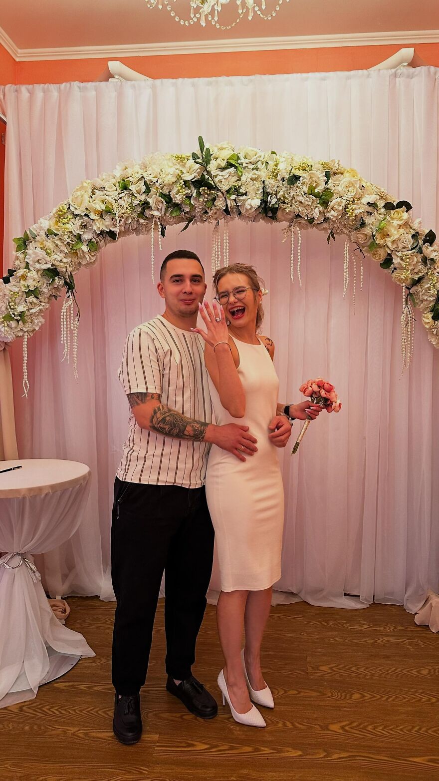 A happy couple showing off a ring under a floral arch, a funny wedding photo of their engagement joy.