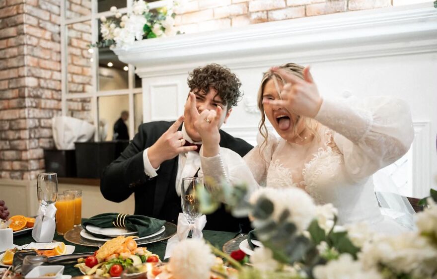 A newlywed couple at their wedding reception making funny faces and rock-and-roll hand gestures, captured in a silly wedding photo.