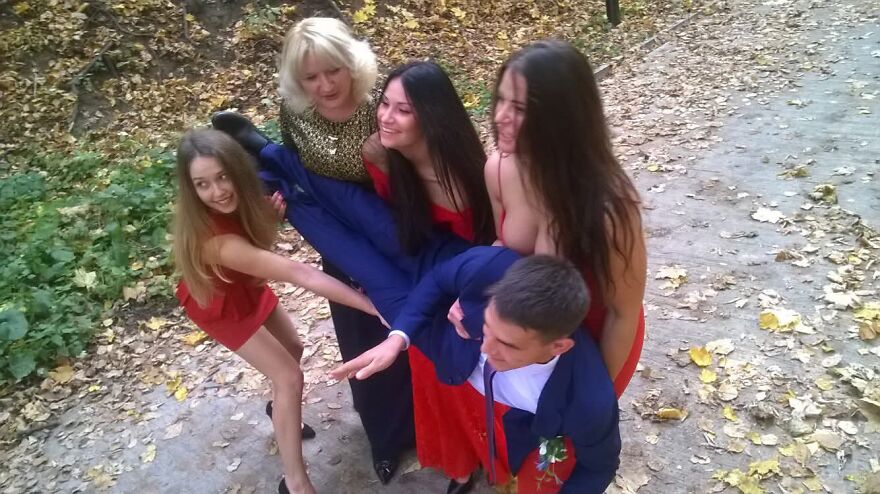 Four women in red dresses humorously lifting a man in a blue suit, a funny wedding photo taken outdoors with autumn leaves.