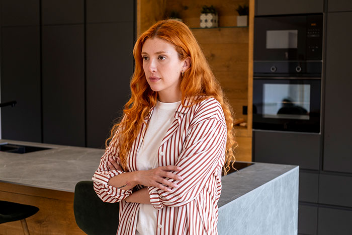 A woman with long red hair, arms crossed, standing in a modern kitchen, considering a college fund apology. A woman with long red hair, arms crossed, standing in a modern kitchen, considering a college fund apology.