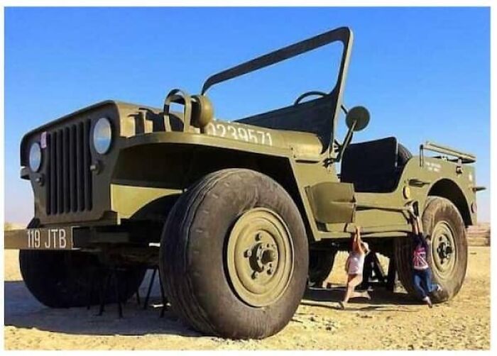 A massive size comparison of a jeep with two people hanging from it, showing the true scale of the vehicle.
