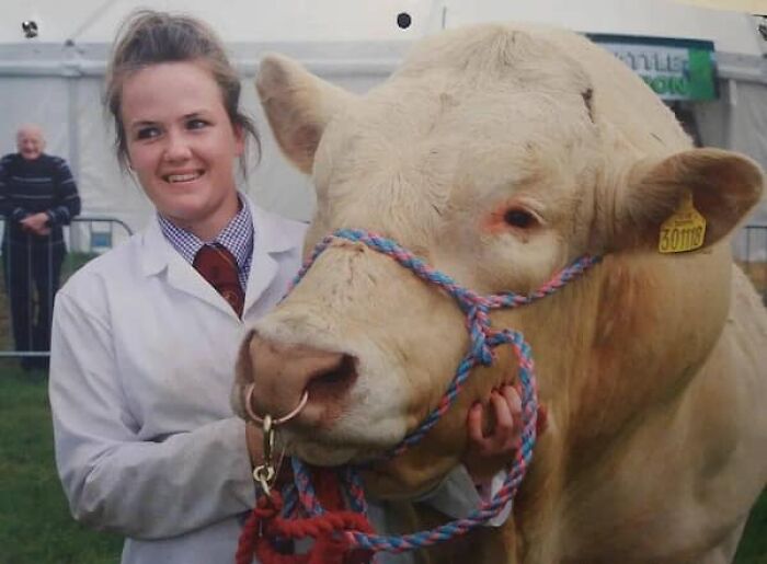 A woman stands proudly next to a large bull, showcasing their true scale in a rural setting.