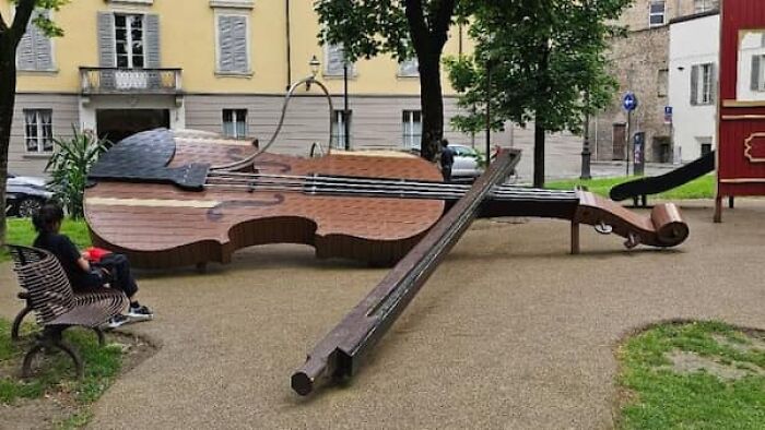 A person sits on a bench next to a giant violin, emphasizing the true scale of the instrument in a park.