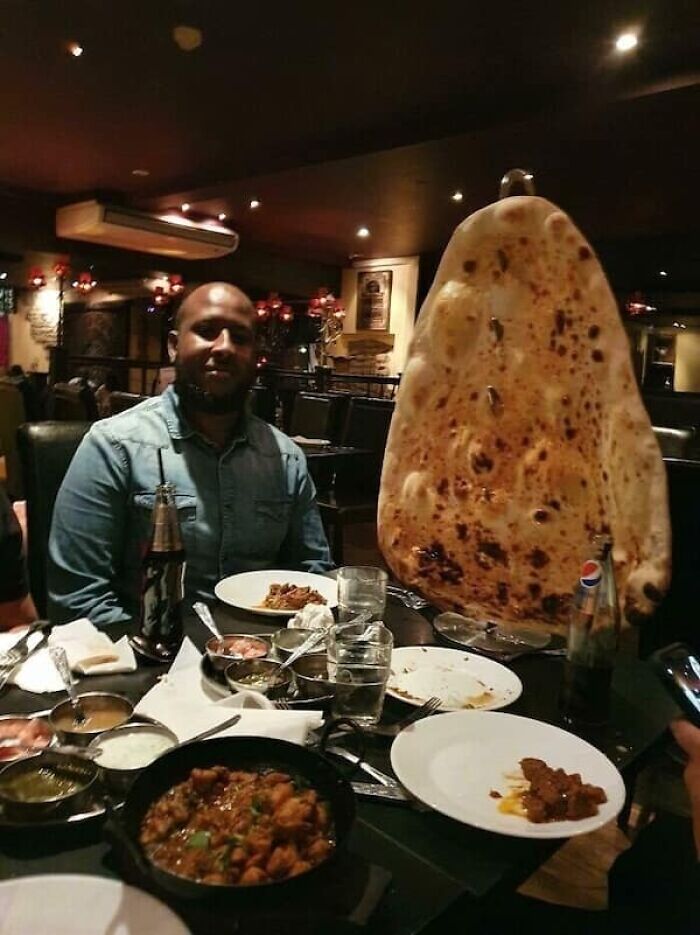 A man smiles behind an enormous naan bread, highlighting the true scale of the food item on a table.