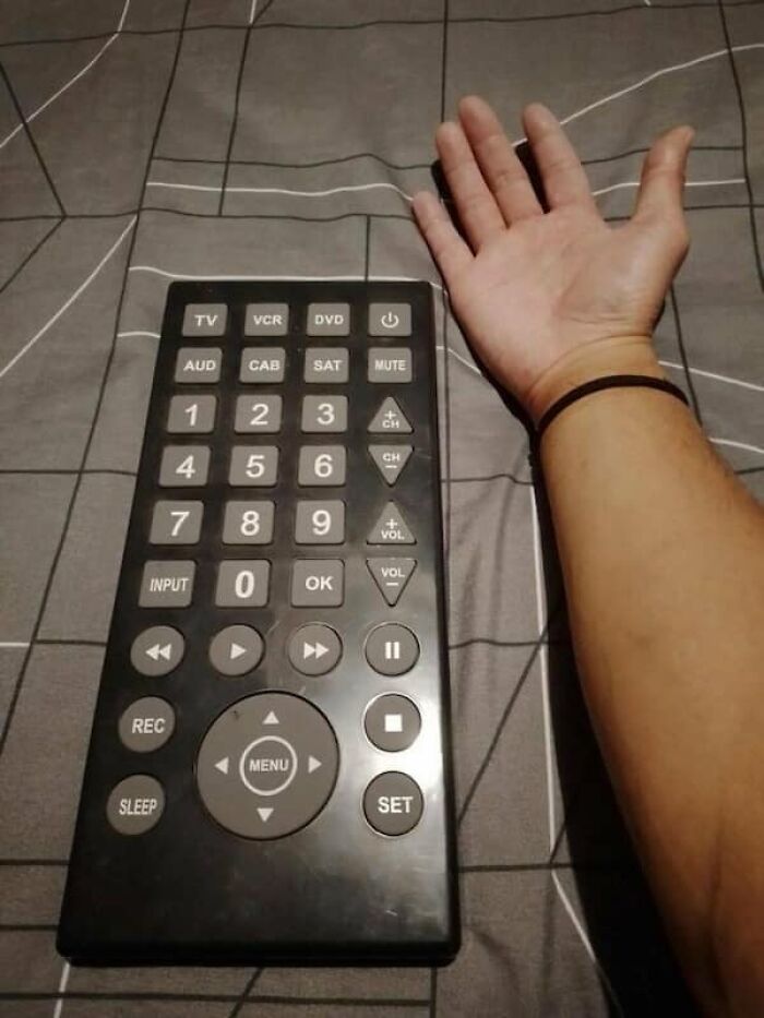 A hand resting next to an oversized remote control, demonstrating the true scale of the device on a bed.
