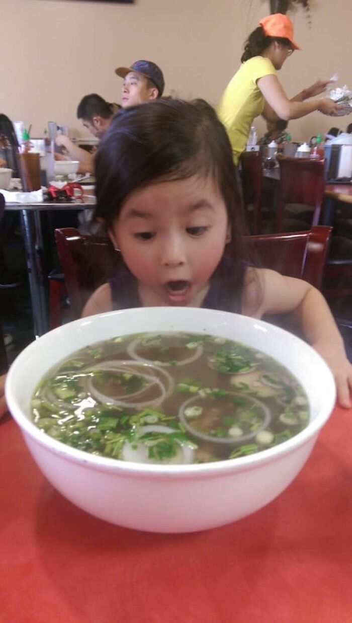 Little girl looking at an enormous bowl of pho with a surprised expression, showing the size comparison of a meal.