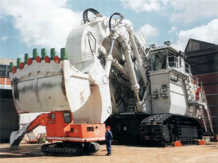 A person stands next to an enormous mining excavator, emphasizing the machine's true scale and immense size.
