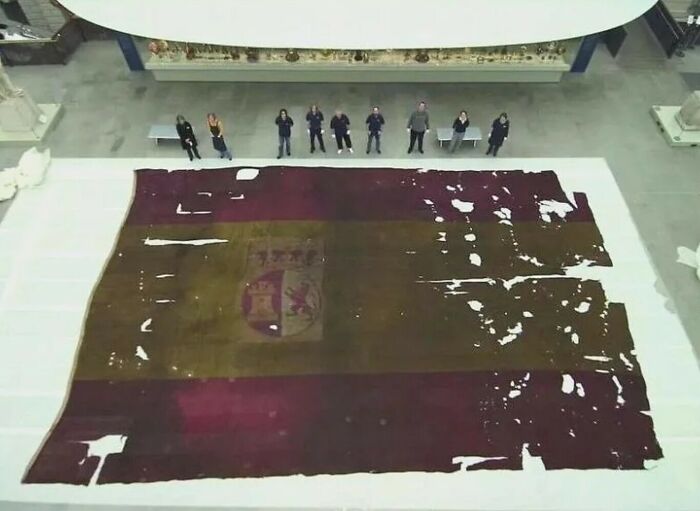 A group of people standing around an enormous, tattered Spanish flag laid on the floor, demonstrating its true scale.