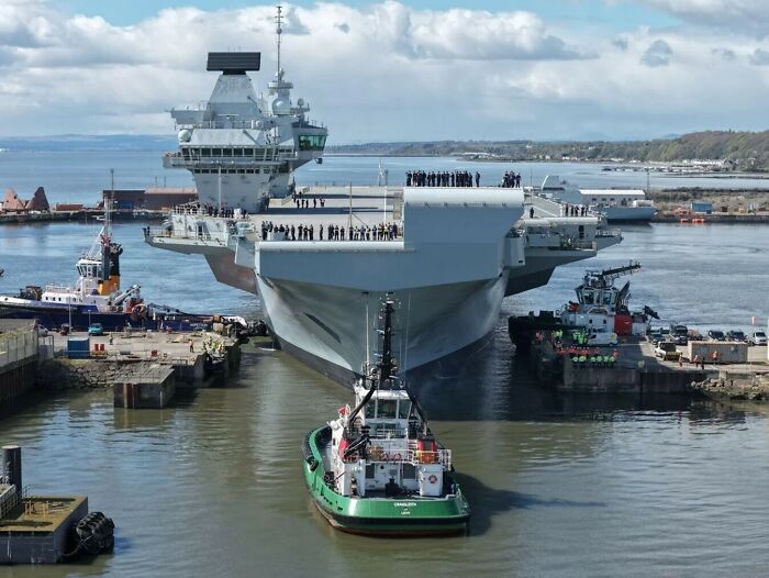 A large aircraft carrier being guided by tugboats in a harbor, demonstrating the true scale of naval vessels.