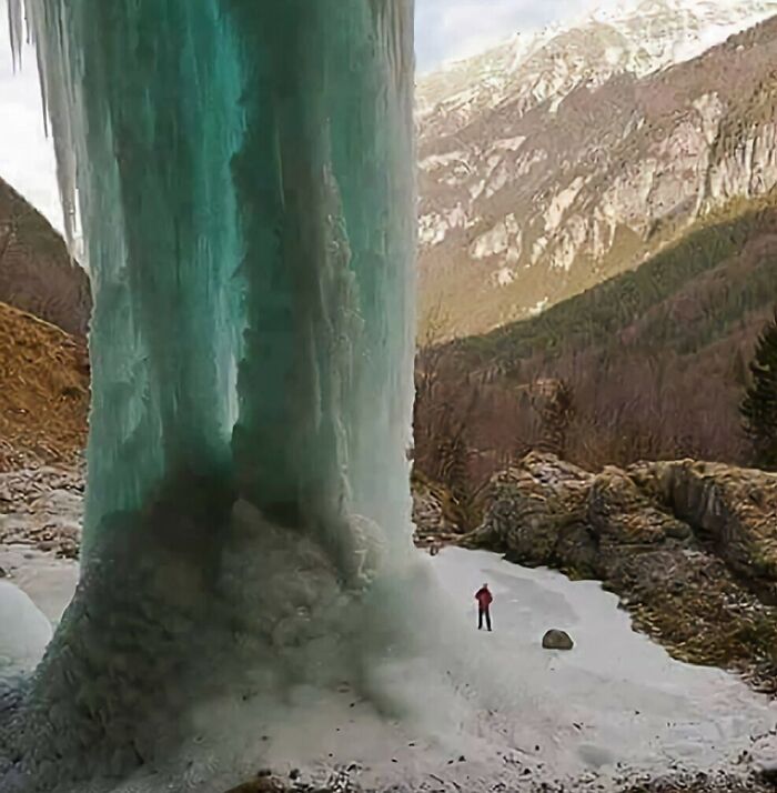 A lone person in red stands at the base of a towering frozen waterfall, illustrating the true scale of the ice formation.