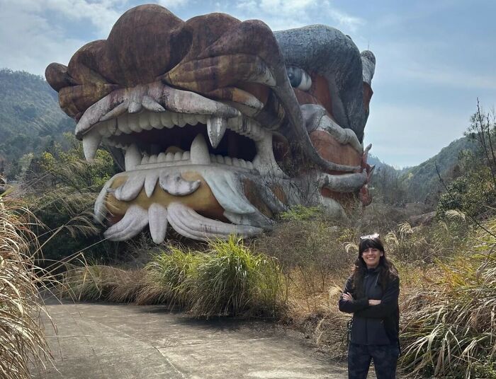 A woman standing in front of a massive, weathered dragon head structure, highlighting the true scale of the sculpture.