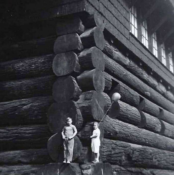Two small children standing in front of a giant log cabin, showcasing the true scale of the logs.